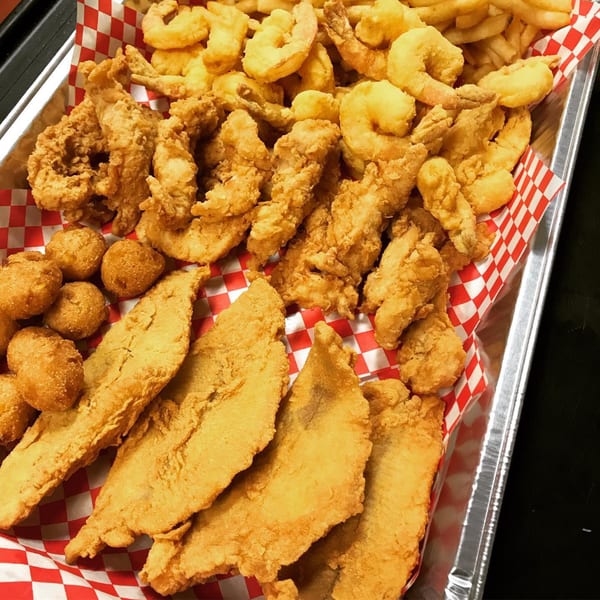 A Louisiana Shack platter with fried shrimp, fish, chicken, hush puppies, and fries in a red checkered basket.